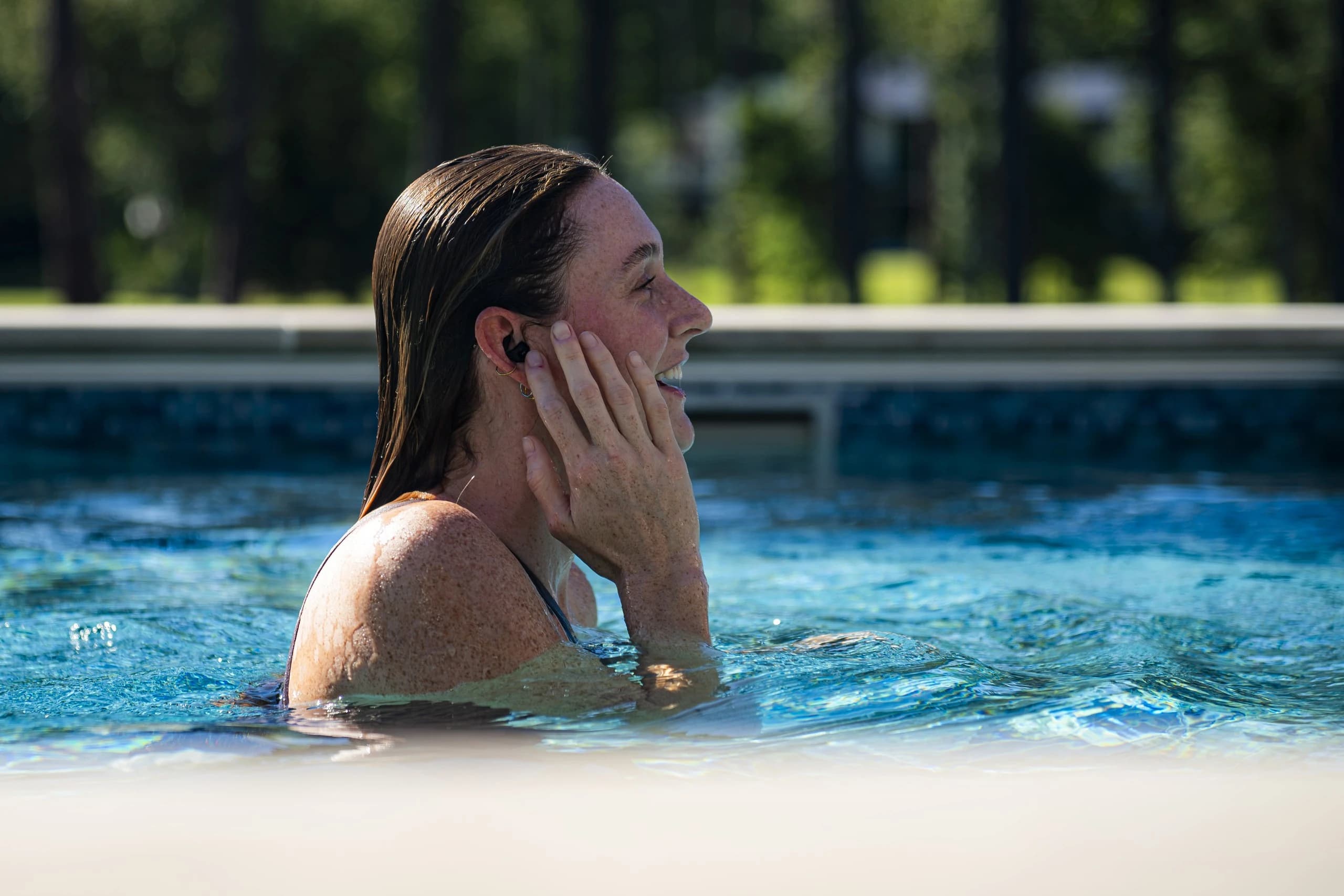 A swimmer in a sunlit pool, smiling, a hand to the SPRTN Swims bud in her ear